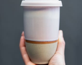 May include: A white ceramic travel mug with a light brown and beige speckled design. The mug has a white lid.
