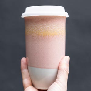 May include: A ceramic travel mug with a white lid. The mug has a pink, yellow, and white speckled design. The mug is cylindrical and appears to be held by a person.