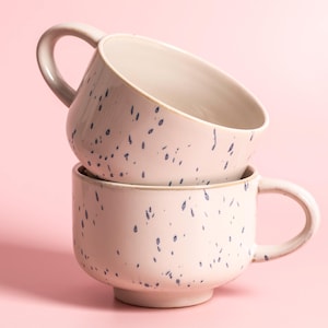 May include: Two stacked, off-white ceramic mugs with a speckled navy blue pattern. Each mug has a rounded handle and a wide, slightly flared rim. The mugs are set against a soft pink background, creating a simple, yet elegant aesthetic.