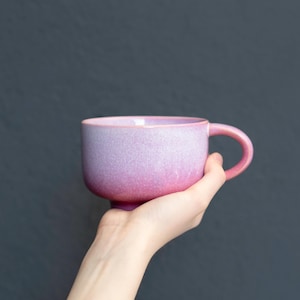 Peut inclure: Une tasse en céramique avec une forme arrondie lisse et une anse incurvée. L'extérieur présente une finition dégradée qui passe d'un lavande pâle et doux en haut à un rose saturé et vibrant à la base, avec une texture mouchetée.