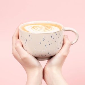 May include: A large, speckled ceramic mug filled with a latte, held by two hands. The latte art features a heart design. The mug is off-white with blue speckles, set against a pink background.