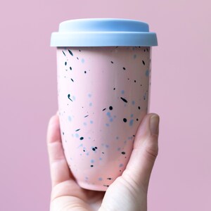 May include: A pink travel mug with a light blue lid. The mug is speckled with blue and black dots. The mug is held in a hand against a pink background. The travel mug is designed to hold approximately 12 ounces of liquid.