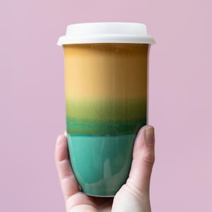 May include: A ceramic travel mug with a white lid. The mug is a gradient of colors, starting with a teal base, transitioning to green, then yellow, and ending with a tan top. The mug is held by a hand.