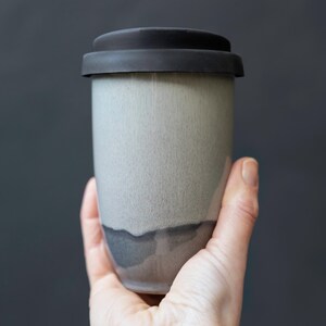 May include: A ceramic travel mug with a matte finish. The mug is primarily light gray with a darker gray band near the bottom and a black lid. The mug is held in a hand against a dark gray background.