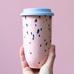 May include: A pink ceramic travel mug with a light blue lid. The mug is decorated with dark blue and gray speckles. The mug is designed to hold hot or cold beverages.