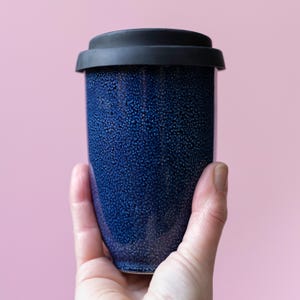 Pode incluir: Uma caneca de viagem azul escuro com uma superfície texturizada e uma tampa preta. A caneca é segurada em uma mão contra um fundo rosa. A caneca de viagem foi projetada para manter as bebidas quentes ou frias.