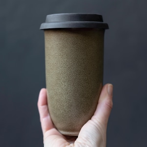 Pode incluir: Uma caneca de viagem de cerâmica verde-oliva salpicada com uma tampa preta. A caneca cilíndrica é segurada em uma mão contra um fundo cinza escuro. A caneca tem aproximadamente 15 cm de altura.