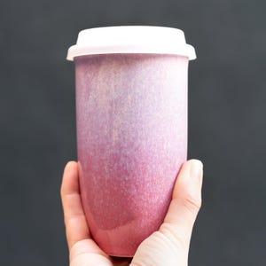 May include: A tall, ceramic travel mug with a white lid. The mug has a gradient color scheme, transitioning from a deep red at the base to a lighter pink near the top. The mug is held in a hand against a dark background.