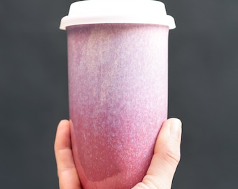 May include: A tall, ceramic travel mug with a white lid. The mug has a gradient color scheme, transitioning from a deep red at the base to a lighter pink near the top. The mug is held in a hand against a dark background.