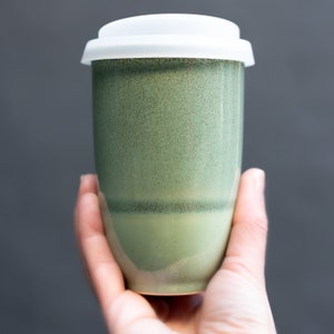 May include: A green ceramic travel mug with a white lid. The mug has a speckled glaze and a light green band around the middle.