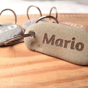 Puede incluir: Primer plano de un llavero de piedra gris con el nombre "Mario" grabado en él. El llavero está unido a un llavero de plata.