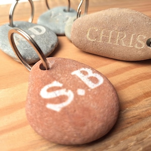 Può includere: Cinque portachiavi in pietra con anelli di metallo. Le pietre sono incise con i nomi "Chris", "S.B.", "A.D." e altri due nomi che non sono visibili nell'immagine.