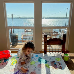 Pode incluir: Uma crian&ccedil;a est&aacute; a colorir uma folha de papel grande com criaturas marinhas. O papel est&aacute; sobre uma mesa em frente a uma janela com vista para um lago. A crian&ccedil;a est&aacute; a usar um marcador roxo para colorir um polvo roxo.