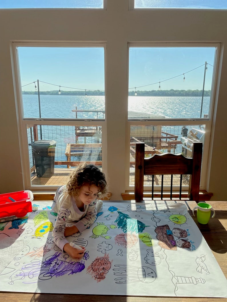 Pode incluir: Uma crian&ccedil;a est&aacute; a colorir uma folha de papel grande com criaturas marinhas. O papel est&aacute; sobre uma mesa em frente a uma janela com vista para um lago. A crian&ccedil;a est&aacute; a usar um marcador roxo para colorir um polvo roxo.