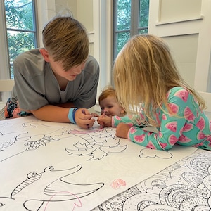May include: Three children are coloring on a large sheet of paper with black and white illustrations of dragons, palm trees, and other designs. The children are using colored pencils and markers to color the illustrations. The paper is on a white table.