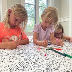 May include: Three children are coloring on a large white tablecloth with black outlines of various shapes and designs. The tablecloth is covered in colorful markers.