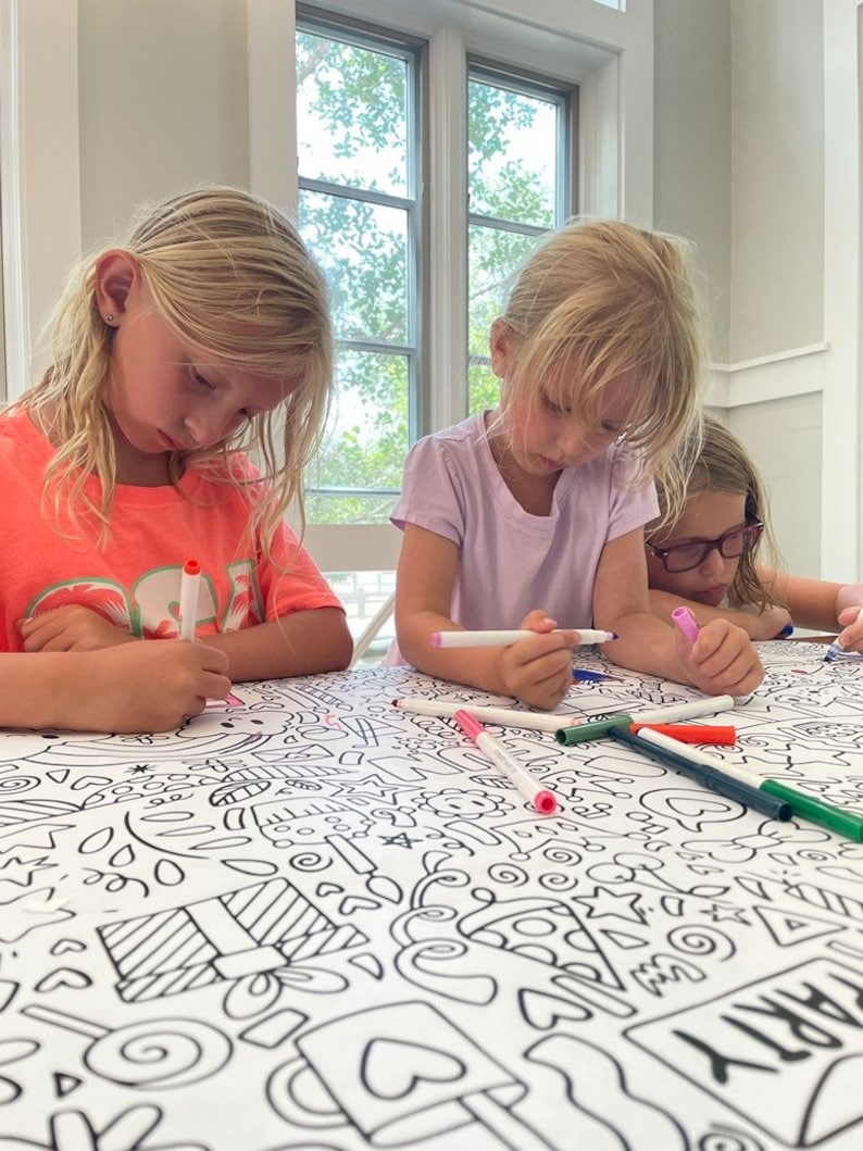 May include: Three children are coloring on a large white tablecloth with black outlines of various shapes and designs. The tablecloth is covered in colorful markers.