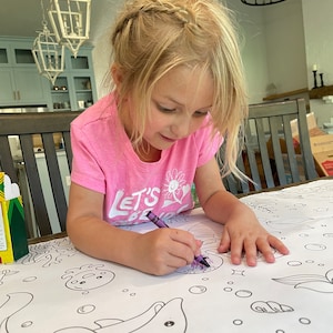 Pode incluir: Uma menina pequena com uma camiseta rosa com o texto "Let's Be Mermaids" est&aacute; colorindo um golfinho grande em uma toalha de mesa branca com um giz de cera roxo. A toalha de mesa tem um design de tema marinho com outras criaturas marinhas e bolhas.