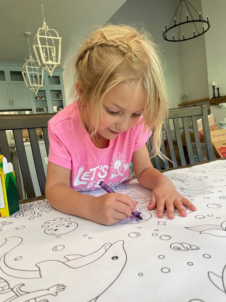 Pode incluir: Uma menina pequena com uma camiseta rosa com o texto "Let's Be Mermaids" est&aacute; colorindo um golfinho grande em uma toalha de mesa branca com um giz de cera roxo. A toalha de mesa tem um design de tema marinho com outras criaturas marinhas e bolhas.