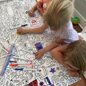May include: Three children color a large white tablecloth with black and white outlines of various objects, including a cake, pizza, and a heart. The tablecloth is covered in colorful markers.