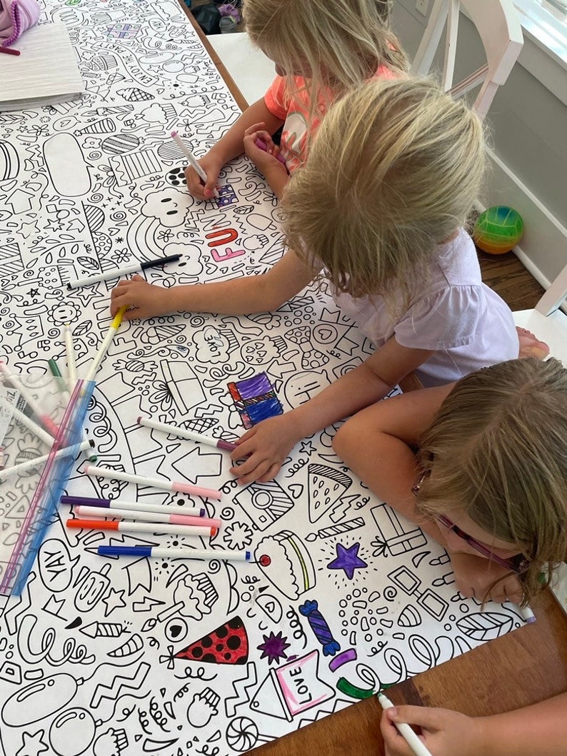 May include: Three children color a large white tablecloth with black and white outlines of various objects, including a cake, pizza, and a heart. The tablecloth is covered in colorful markers.