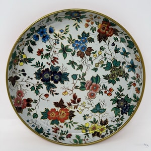 Puede incluir: Una bandeja redonda de metal con base blanca que presenta un patrón floral detallado en tonos azul, rojo, amarillo y verde. La bandeja tiene un borde fino y elevado de color dorado que rodea toda la circunferencia del plato circular.