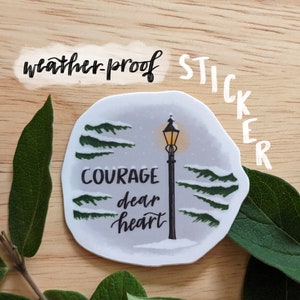 May include: A weather-proof sticker featuring a winter scene with a lamppost and the words "Courage dear heart." The sticker has a light gray background with green trees and snow. The word "sticker" is written vertically.