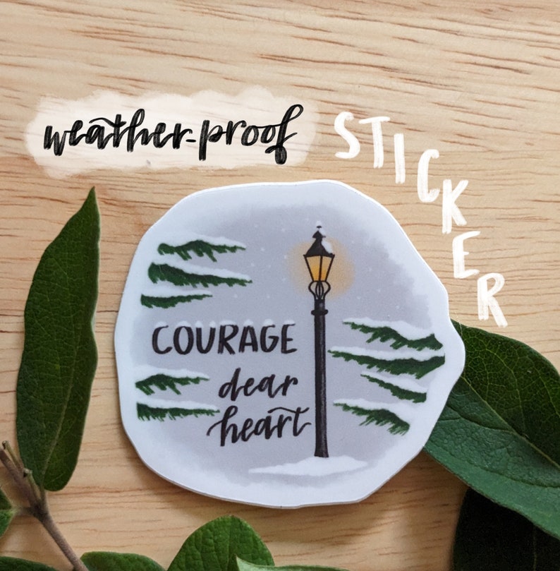 May include: A weather-proof sticker featuring a winter scene with a lamppost and the words "Courage dear heart." The sticker has a light gray background with green trees and snow. The word "sticker" is written vertically.