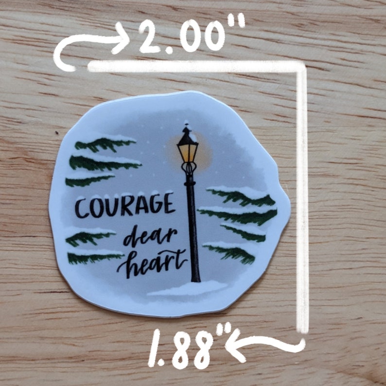 May include: A die-cut sticker with a winter scene featuring a lamppost and the words "Courage dear heart." The sticker is approximately 2.00" x 1.88" and has a snowy background with evergreen trees.