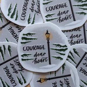 May include: A collection of stickers featuring a winter scene with a lamppost and evergreen trees. The text "COURAGE dear heart" is written in a script font. The stickers have a white border and a light gray background.