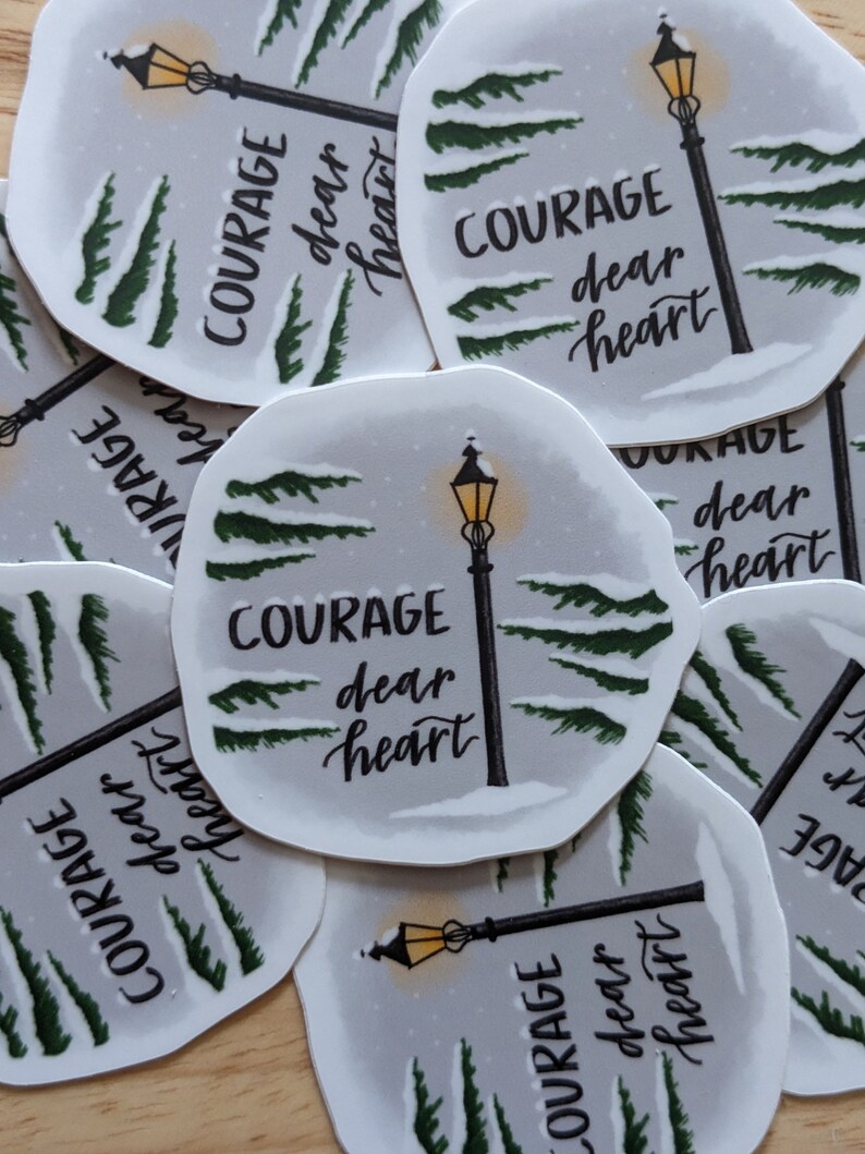 May include: A collection of stickers featuring a winter scene with a lamppost and evergreen trees. The text "COURAGE dear heart" is written in a script font. The stickers have a white border and a light gray background.