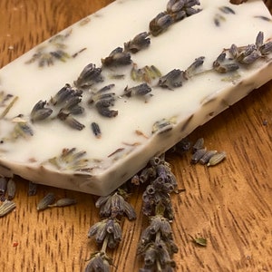 May include: A white wax melt with dried lavender flowers embedded in it. The wax melt is shaped like a tag and has a brown leather cord attached to it.
