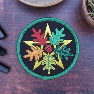 May include: Embroidered patch with a black background, a yellow star, and colorful leaves in red, green, and orange. The patch has a green border and an acorn in the center. The patch is round.