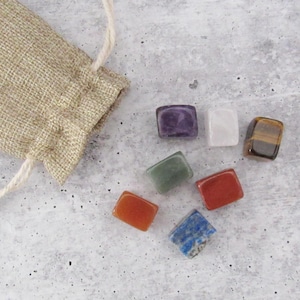 May include: Seven small, square-shaped crystals in various colours, including purple, white, brown, green, red, blue, and orange, are arranged on a grey surface. The crystals are in a small hessian bag.