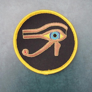 May include: A circular embroidered patch featuring the Eye of Horus symbol in copper-colored thread on a black background. The eye has a bright blue iris and a small red dot at the end of the tail. The patch is finished with a thick yellow border.