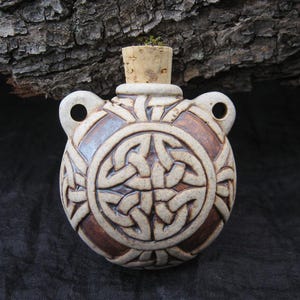 May include: A ceramic flask with a cork stopper. The flask is round with a brown and cream-colored Celtic knot design. It has two small loop handles on either side of the top.