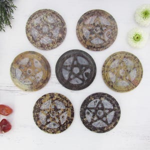 Pode incluir: Sete porta-copos circulares de pedra dispostos em grupo, cada um com um design de pentagrama esculpido. Os porta-copos exibem tons variados de castanho, bege e cinzento com superfícies polidas e texturas de pedra.