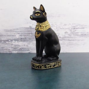 Puede incluir: Figura de gato egipcio negro con detalles dorados. El gato está sentado sobre una base con jeroglíficos.