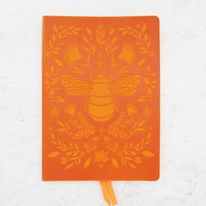 May include: Orange journal with a bee and floral design. The cover features a large bee in the center, surrounded by detailed floral and leaf patterns. A matching orange ribbon bookmark is attached.
