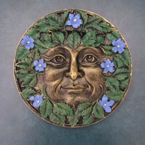 Peut inclure: Plaque murale ronde de couleur bronze représentant un visage souriant orné d'une couronne de feuilles vertes et de fleurs bleues. Le visage présente des détails et un bord doré. La plaque est probablement en résine ou dans un matériau similaire.
