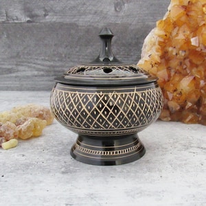 Puede incluir: Quemador de incienso negro y dorado con un patrón de diamante decorativo. El quemador tiene una tapa con agujeros para quemar incienso.