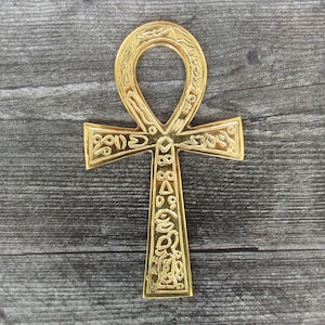 Könnte beinhalten: Ein goldfarbener Ankh-Anhänger mit filigranen Details und Hieroglyphen. Der Ankh ist ein Symbol für Leben und ewiges Leben in der alten ägyptischen Mythologie.