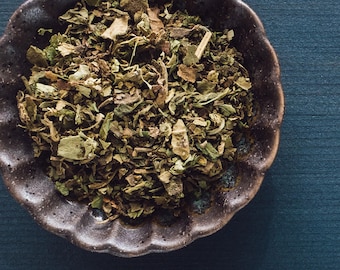 May include: A close-up of a small brown bowl filled with dried herbs. The bowl has a scalloped edge and is sitting on a dark blue surface.
