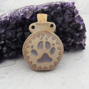 May include: A small ceramic bottle with a cork stopper. The bottle is decorated with a paw print and a geometric pattern. The bottle is a light brown colour with a hint of purple.