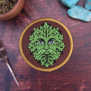 May include: Round embroidered patch featuring a green Green Man face with a leafy beard and mustache, set against a brown background. The patch has a gold border and is likely for clothing or accessories.