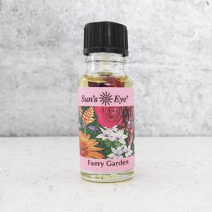Puede incluir: Una pequeña botella de vidrio de aceite esencial de jardín de hadas con una tapa negra. La botella tiene una etiqueta rosa con las palabras "Sun's Eye" y "Faery Garden" impresas en ella. La etiqueta también presenta una ilustración colorida de flores.