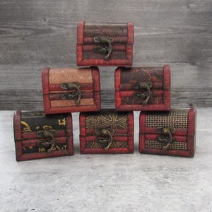 Może przedstawiać: Sześć małych drewnianych skrzyń ze skarbami z ozdobnymi wzorami i metalowymi klamrami. Skrzynie są ułożone w kształt piramidy na szarej powierzchni.