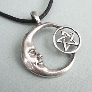 May include: Silver-toned pendant featuring a crescent moon with a face and a five-pointed star within a circle. The pendant hangs from a black cord. The design has a celestial and mystical theme.