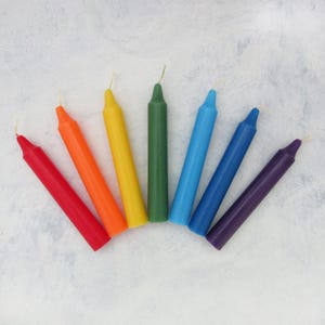 May include: A set of seven thin, taper candles in the colors of the rainbow. The candles are arranged in a semi-circle, with the red candle at the bottom left and the purple candle at the bottom right.