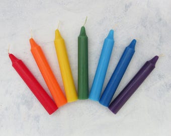 Könnte beinhalten: Ein Satz von sieben dünnen, spitz zulaufenden Kerzen in den Farben des Regenbogens. Die Kerzen sind in einem Halbkreis angeordnet, wobei die rote Kerze unten links und die violette Kerze unten rechts liegt.